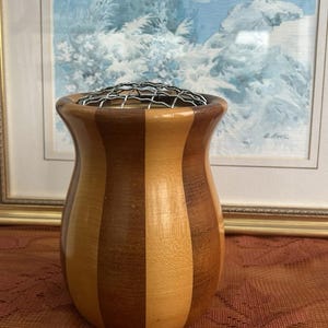 May include: A decorative wooden vase with vertical stripes in light and dark brown. The vase has a bulbous shape and a metal grid at the top. The background features a framed painting with a landscape scene.