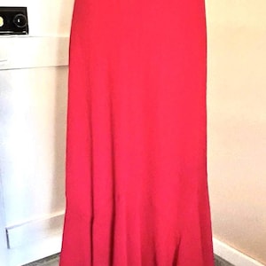 May include: A long, flowing red skirt on a mannequin. The skirt is a solid, vibrant red color and has a slight flare at the bottom. The fabric appears to be lightweight and has a subtle sheen.