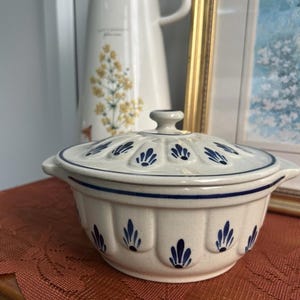 Könnte beinhalten: Cremefarbene Keramik-Auflaufform mit Deckel, mit blauen floralen Designs und einem blauen Rand. Die Schüssel hat zwei Griffe. Eine weiße Kanne mit gelben floralen Designs befindet sich im Hintergrund. Die Schüssel steht auf einer braunen Oberfläche.