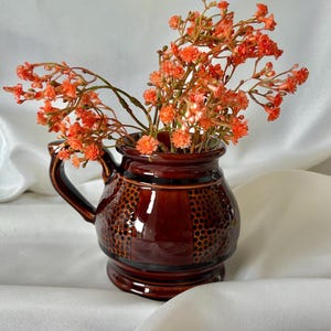 May include: A small, glossy dark brown ceramic jug with a handle and a textured, dotted pattern around the midsection. The jug is filled with a cluster of vibrant orange baby's breath flowers, which extend outwards from the opening.