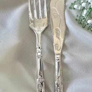 May include: A silver-toned metal fork and knife set featuring ornate, embossed floral and shell-like detailing on the handles. The knife blade displays a curved, etched leaf pattern, while the fork has four tines and a matching decorative base.