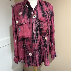 Puede incluir: Una camisa de manga larga con un diseño tie-dye en tonos rosa y negro. La camisa tiene dos bolsillos en el pecho y pequeños adornos coloridos. La camisa se exhibe en un maniquí.