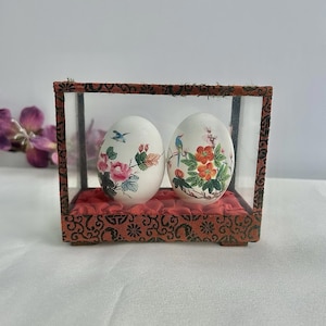 May include: Two decorative eggs with floral and bird designs are displayed inside a glass case with a red patterned base. The case has a dark brown frame with an ornate design. The eggs are white with colourful illustrations.