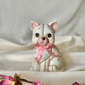 May include: A ceramic figurine of a seated white dog with large dark eyes and long eyelashes. The dog features a delicate floral pattern, pink-spotted ears, and a pink satin ribbon tied around its neck. The surface has subtle gold-toned outlines.