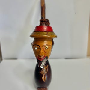 May include: A hand-carved wooden bottle stopper featuring a stylized face with a red hat, a dark brown body, and a small silver piece. The stopper has a rope detail and a carved tulip design.