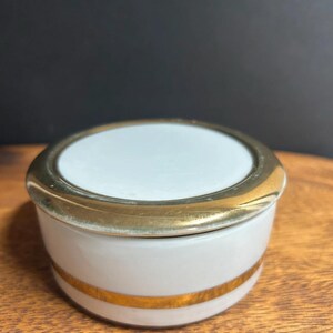 May include: A small, round, white ceramic trinket box with a gold-colored band and trim. The lid is white with a gold-colored rim. The box is sitting on a wooden surface.
