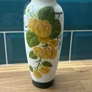 Puede incluir: Un jarrón alto de cerámica blanquecina con base y borde negros. El jarrón está decorado con racimos de bayas amarillas, hojas verdes y pequeñas flores azules y blancas. El jarrón mide unos 25 cm de alto.