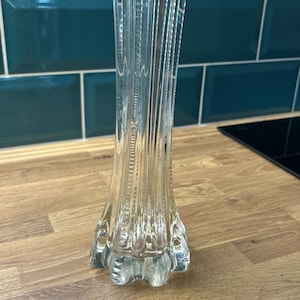 Pode incluir: Um vaso de vidro transparente com um design canelado. O vaso tem uma base alargada e um pescoço alto e esguio. O vaso está sobre uma superfície de madeira, com um fundo de azulejos verde-azulado.