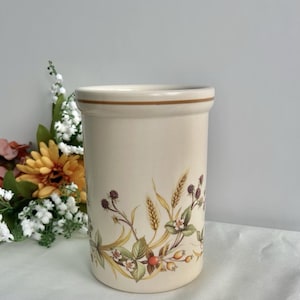 May include: Cream-coloured ceramic utensil holder with a brown rim and a floral design. The design features wheat, berries, and leaves in shades of green, brown, and yellow. The holder is cylindrical and stands approximately 15 cm tall.