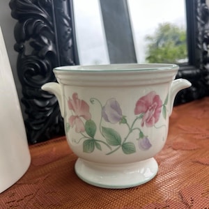 Puede incluir: Una maceta de cerámica blanca con base de pedestal y dos asas pequeñas. La maceta está decorada con motivos florales rosas y morados y hojas verdes. Un ribete verde claro acentúa la parte superior e inferior.