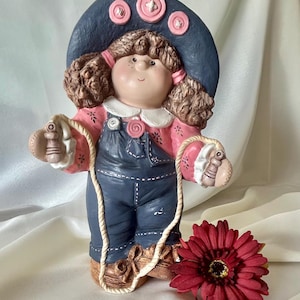 Può includere: Una statuetta in ceramica di un bambino con capelli castani ricci, che indossa un cappello blu scuro con tre bottoni rosa, una camicia rosa a maniche lunghe con motivi floreali e una salopette blu scuro. La figura tiene una corda per saltare vicino a un fiore rosso.