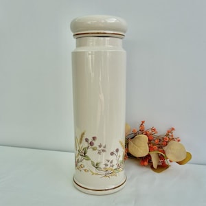 May include: A cream-coloured ceramic jar with a lid, featuring a botanical design of berries and leaves. The jar has a glossy finish and brown detailing. Dried flowers are placed beside it.
