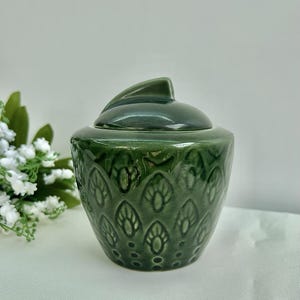 May include: A green ceramic jar with a lid. The jar has a textured, repeating pattern of leaf-like shapes. The lid has a fin-like handle. The jar is a deep, forest green color.