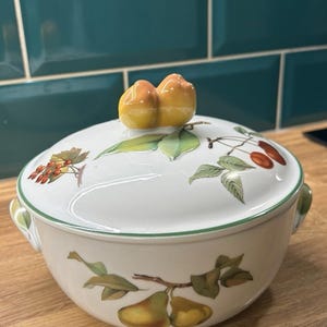 Pode incluir: Tigela de cerâmica branca com tampa, com um design de frutas e folhas. A tampa é encimada por um par de frutas amarelo-alaranjadas. A tigela tem uma borda e alças verdes. As ilustrações de frutas incluem peras e cerejas.