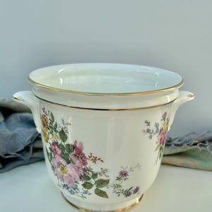 Puede incluir: Hielera de cerámica blanca con tapa, adornada con un estampado floral en rosa, amarillo y azul. El borde y las asas están acentuados con ribetes dorados. Una pieza decorativa para servir.