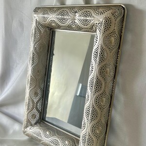 May include: A rectangular mirror featuring a silver-toned metal frame with an intricate, perforated lattice pattern. The frame displays a repeating wave-like design with small, smooth circular accents embedded throughout the metallic surface.