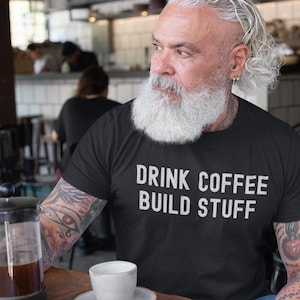 Könnte beinhalten: Ein Mann trägt ein schwarzes T-Shirt mit dem Text "DRINK COFFEE BUILD STUFF" in weißen Buchstaben. Er hält eine French Press Kaffeekanne und eine kleine weiße Tasse.