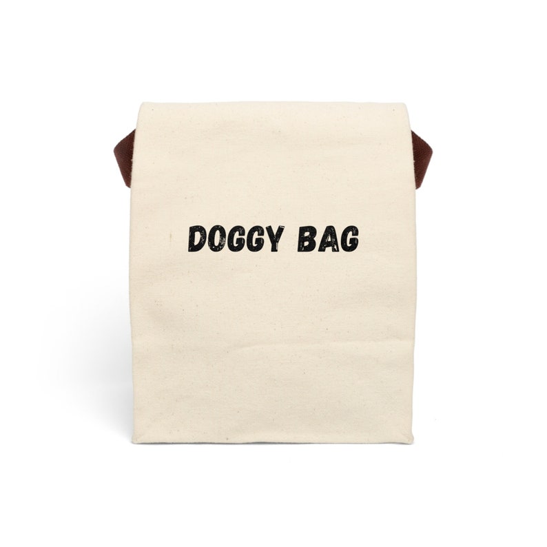 Może przedstawiać: Biała torba z pł&oacute;tna z brązowymi sk&oacute;rzanymi uchwytami i nadrukiem "DOGGY BAG" w kolorze czarnym.