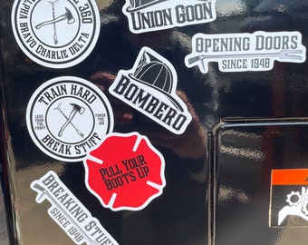 May include: A collection of firefighter-themed stickers with various designs and text. The stickers feature images of axes, helmets, and the text "Union Goon," "Bombero," "Opening Doors Since 1948," and "Pull Your Boots Up." The stickers are primarily black, white, and red.