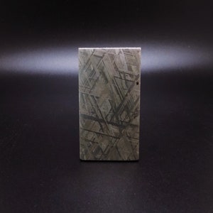 May include: A rectangular, gray-toned meteorite with a Widmanstätten pattern. The iron meteorite displays a geometric crystalline structure, with intersecting lines and shapes. The object is set against a dark background.