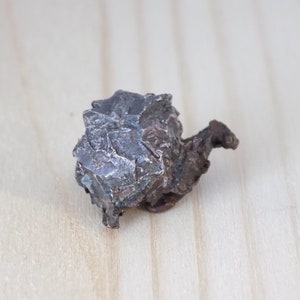 May include: A dark, irregularly shaped rock with a rough, textured surface. The rock has a metallic appearance and sits on a light-colored wooden surface. The object appears to be a mineral specimen.
