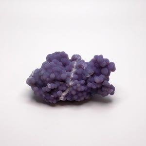 Puede incluir: Un grupo de piedra preciosa de ágata de uva púrpura, con una textura botrioidal y burbujeante. La piedra tiene un tono púrpura claro con vetas blancas. La piedra preciosa está sobre un fondo blanco.