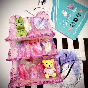 May include: A pink three-tiered shelf with a bear and bunny figurine. The shelf is decorated with glitter, moons, and pink and white swirls. The shelf is made of resin and is a great way to display small items.