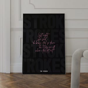 May include: Black framed poster with the band name "STROKES" in a large, bold font. The poster features lyrics in a pink script font, with the band's name at the bottom. The poster is displayed against a white wall and wooden floor.