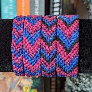 May include: Four friendship bracelets with a chevron pattern in pink, blue, and black colors. The bracelets are stacked on top of each other and are displayed on a black surface.