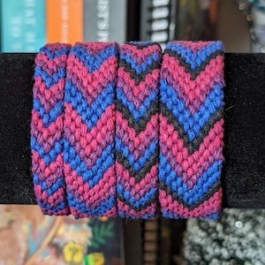 May include: Three friendship bracelets with a chevron pattern in shades of pink, blue, and black.