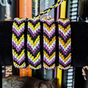 May include: Four friendship bracelets in yellow, black, white, and purple with a chevron pattern. The bracelets are displayed on a black surface in front of a bookshelf.