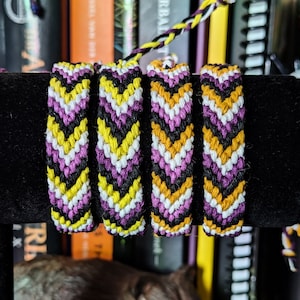 May include: Four friendship bracelets in yellow, black, white, and purple with a chevron pattern. The bracelets are displayed on a black surface in front of a bookshelf.