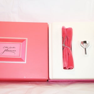May include: A pink gift box set with the words "Estee Lauder pleasures" in a pink and silver frame. Inside, a silver perfume bottle with pink gems, a small silver funnel, and a pink velvet accessory are displayed.