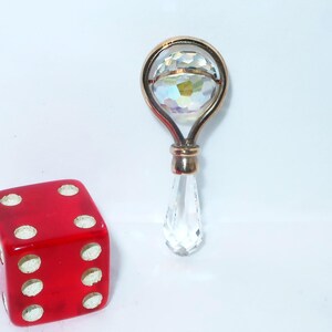 May include: A red six-sided die with white pips positioned next to a gold-toned metal pendant featuring a spherical faceted crystal set within a circular frame and a dangling clear teardrop-shaped crystal prism at the base.