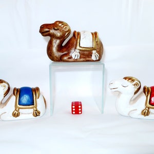 May include: A collection of ceramic camel figurines in various colors. One brown camel sits atop a glass shelf, while two white camels with blue and red saddles are positioned below. A red die with white dots is in the center.