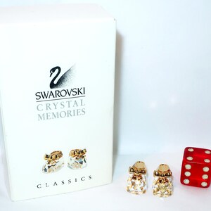 Puede incluir: Una caja blanca de Swarovski Crystal Memories Classics con un logotipo de cisne y texto, acompañada de dos figuras de zapatitos de bebé de cristal transparente con lazos dorados y un dado rojo translúcido de seis caras con puntos blancos.