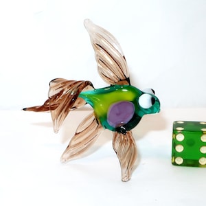 May include: A colorful glass fish figurine with brown fins and tail, and a green and purple body. A green die with white dots is next to the fish. The fish is approximately 4 inches long.
