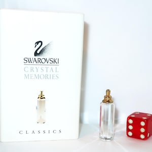 Puede incluir: Una caja rectangular blanca de Swarovski Crystal Memories Classics con un logotipo de cisne negro y texto, junto a una botella miniatura de cristal facetado transparente con tapa dorada y un dado rojo pequeño con puntos blancos.