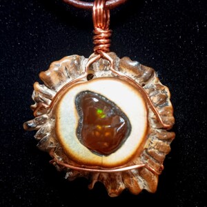 May include: A handcrafted pendant featuring a polished, brown gemstone set within a cream-colored, antler-shaped frame. The pendant is wrapped with copper wire and hangs from a brown leather cord. The gemstone has a green highlight.