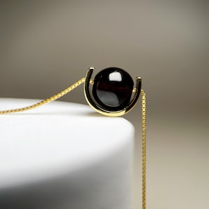 May include: A gold chain necklace with a dark red gemstone pendant. The pendant is set in a gold frame that resembles a horseshoe.