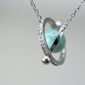 Puede incluir: Un collar de plata con un colgante de piedra turquesa giratorio rodeado de un círculo de pequeños diamantes.