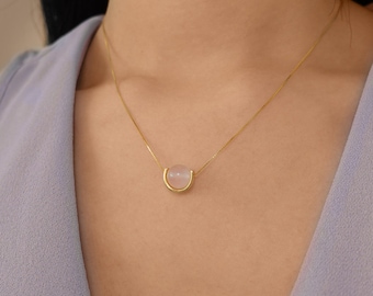 May include: A gold chain necklace with a delicate, curved pendant featuring a round, light pink gemstone.