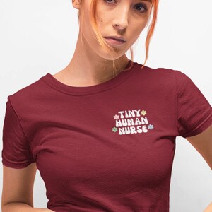 Op de afbeelding: Een vrouw draagt een bordeauxrood t-shirt met de tekst "TINY HUMAN NURSE" in wit met kleurrijke bloemen rond de tekst. Ze draagt een zwarte denim rok met knoopdetails.