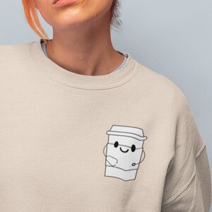 May include: A light beige sweatshirt with a cartoon coffee cup graphic on the left chest. The coffee cup has a smiling face and is holding a small cup.