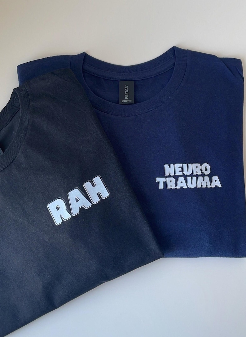 May include: Two t-shirts, one black and one navy blue, with white text. The black t-shirt has the word "RAH" printed on it. The navy blue t-shirt has the words "NEURO TRAUMA" printed on it.