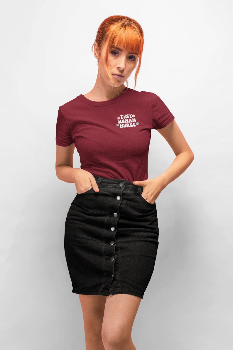 Op de afbeelding: Een vrouw draagt een bordeauxrood t-shirt met de tekst "TINY HUMAN NURSE" in wit met kleurrijke bloemen rond de tekst. Ze draagt een zwarte denim rok met knoopdetails.