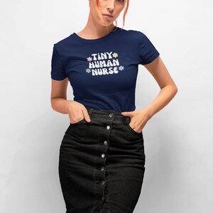 Op de afbeelding: Een marineblauw t-shirt met de tekst "TINY HUMAN NURSE" in wit met pastelkleurige bloemen rond de tekst.