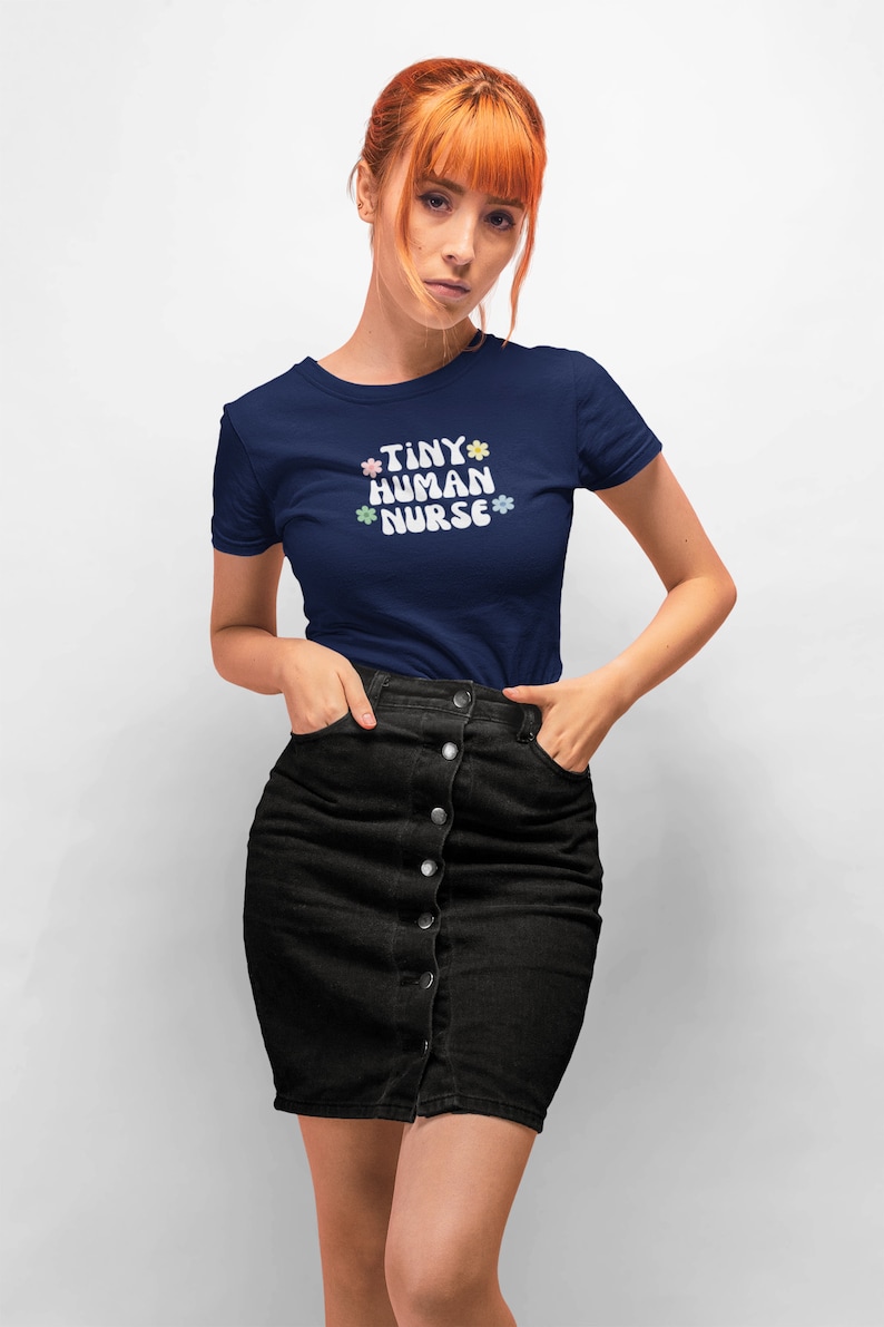 Op de afbeelding: Een marineblauw t-shirt met de tekst "TINY HUMAN NURSE" in wit met pastelkleurige bloemen rond de tekst.