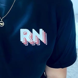 May include: Black t-shirt featuring the letters "RN" in a layered, 3D design with white and pink accents. The shirt is a solid color and appears to be made of a soft material. A gold necklace is visible.