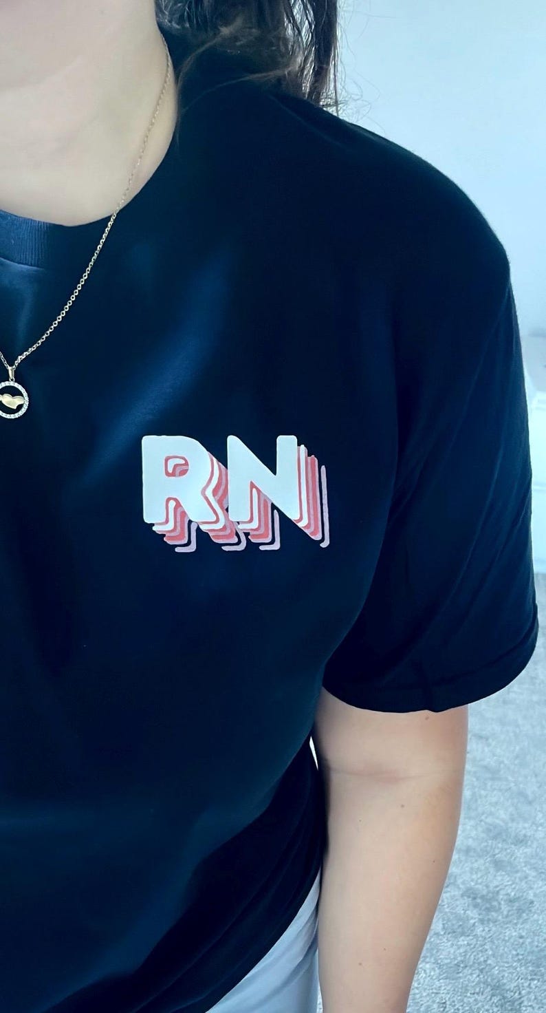May include: Black t-shirt featuring the letters "RN" in a layered, 3D design with white and pink accents. The shirt is a solid color and appears to be made of a soft material. A gold necklace is visible.